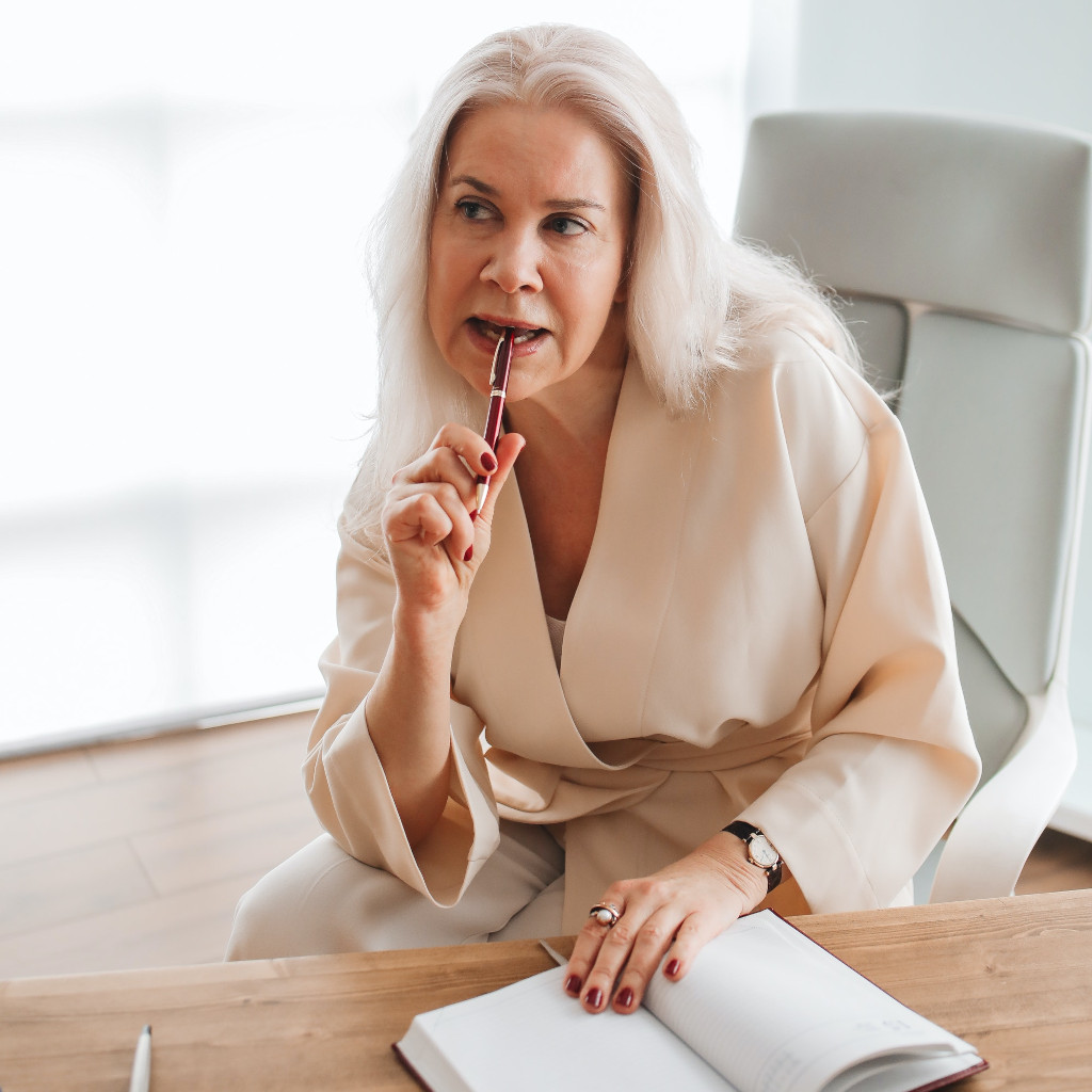JOURNALLING for stress relief - A woman over 50 with long blonde hair sits in a modern office chair beside a desk with an open journal. She is holding a Parker pen and pondering what to write.