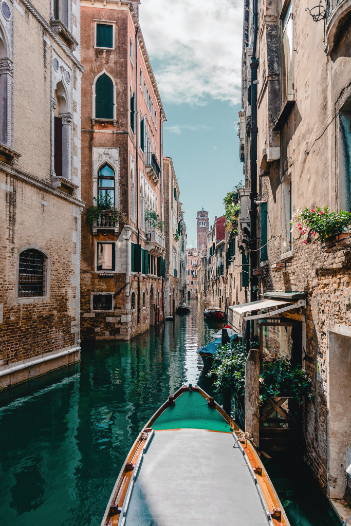 The front of a canal boat moves through the deep green water of a Venice canal between tall warm coloured brick walls, it feels serene