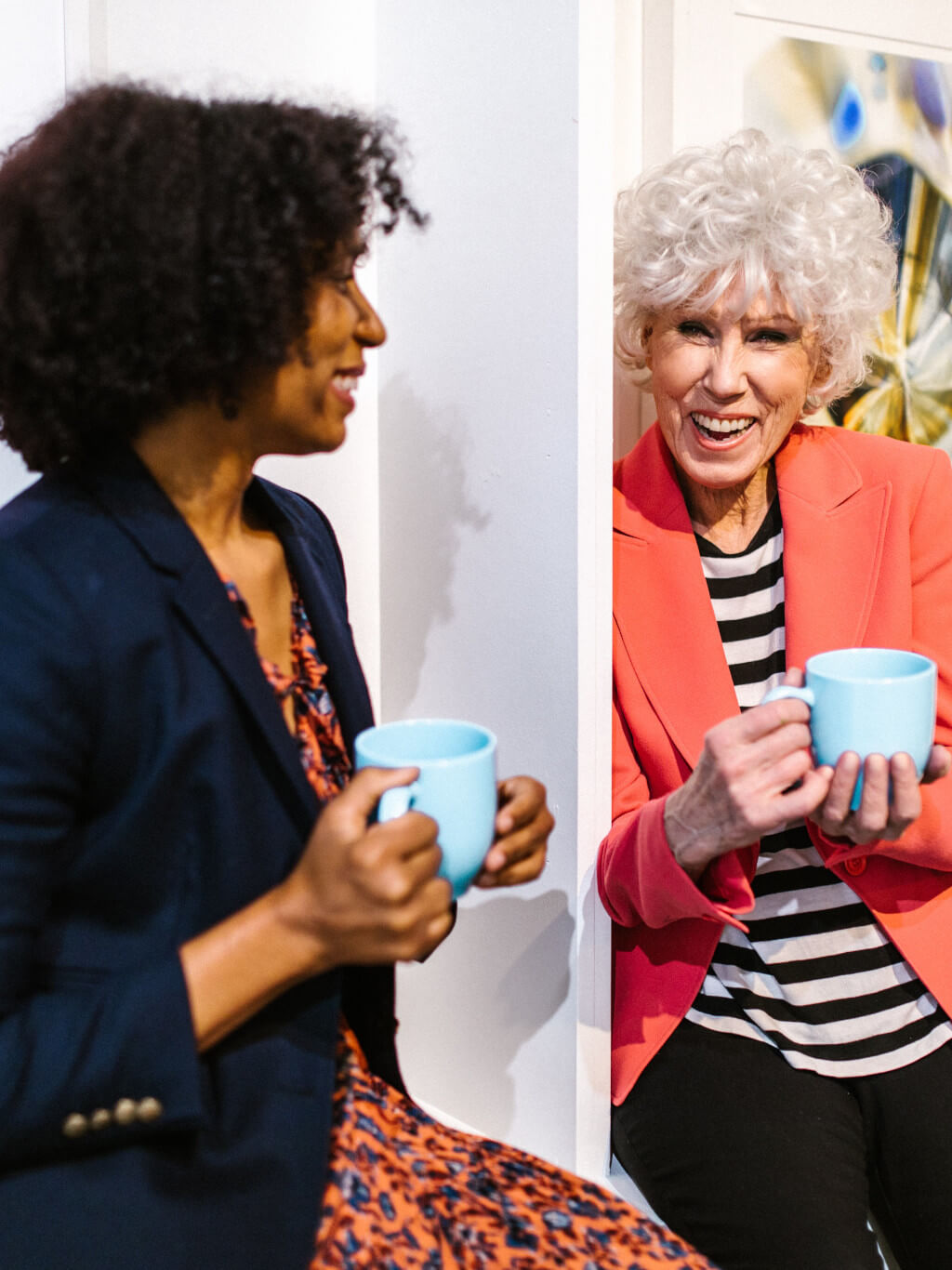 Two women over 50 sit laughing as they have a coffee break together. One woman has a striped black and white t-shirt and a coral jacket and she has curly white hair. The other woman has a dark brown curly bob and is wearing a flowery dress with a blue jacket